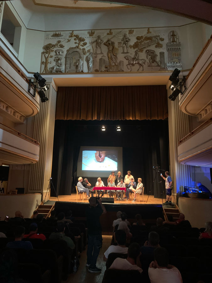 Sala del teatro Titano con la vista dal pubblico e il palco sul fondo, con gli ospiti attorno ad un tavolo alle cui spalle appare una proiezione su grande schermo